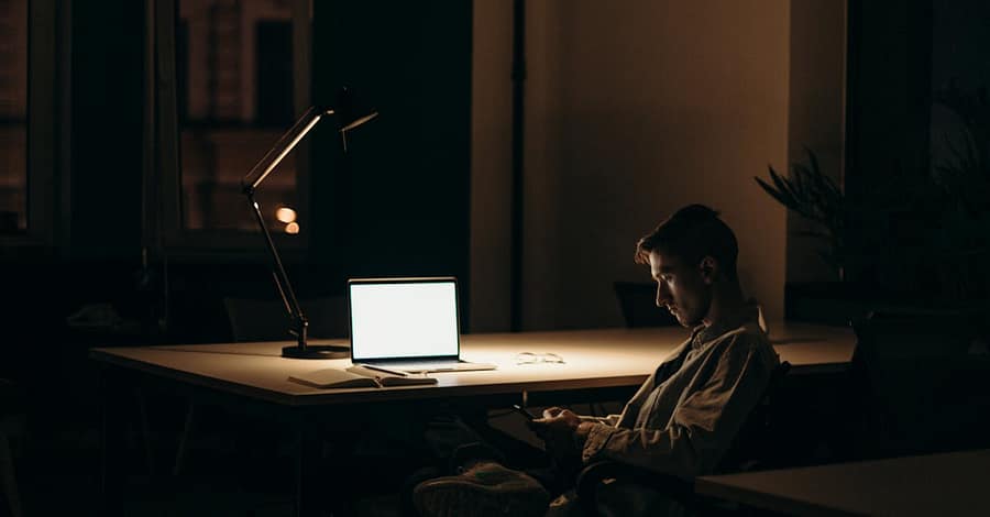 A lone man working on a laptop under dim lighting in a quiet office at night.